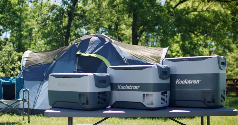 Picture of a campsite with Koolatron's SK30 30 liter capacity, SK40 40 liter capacity, and SK50 50 liter capacity cool boxes on a folding table in the foreground and a dome tent, folding camping chair, and trees in the background