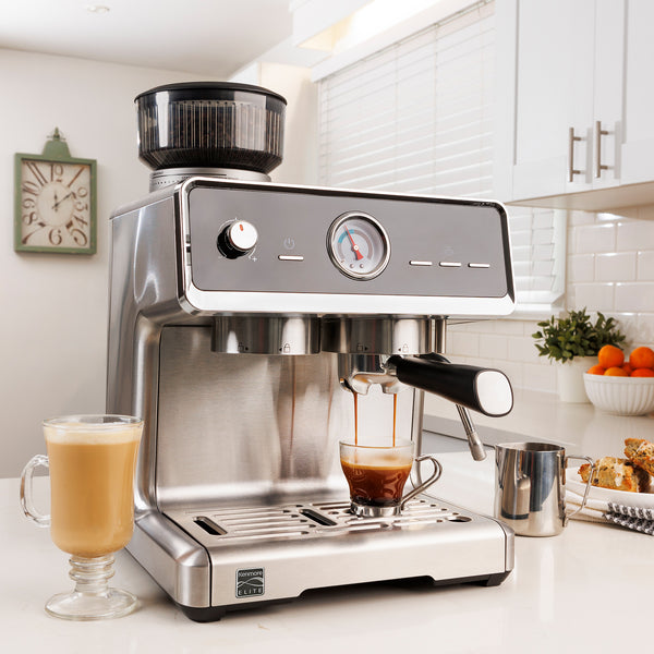 Kenmore Elite auto-tamp espresso maker with espresso being extracted into a clear glass cup and a latte in a clear glass mug to the left on a white counter in a bright clean kitchen